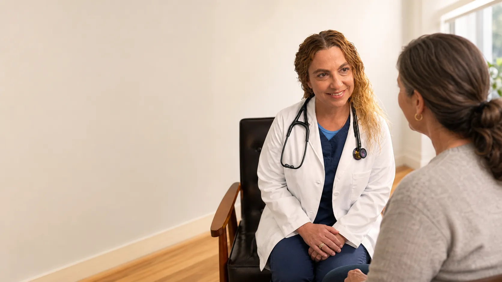 A nurse practitioner in a white coat sitting and talking with a patient in a bright exam room